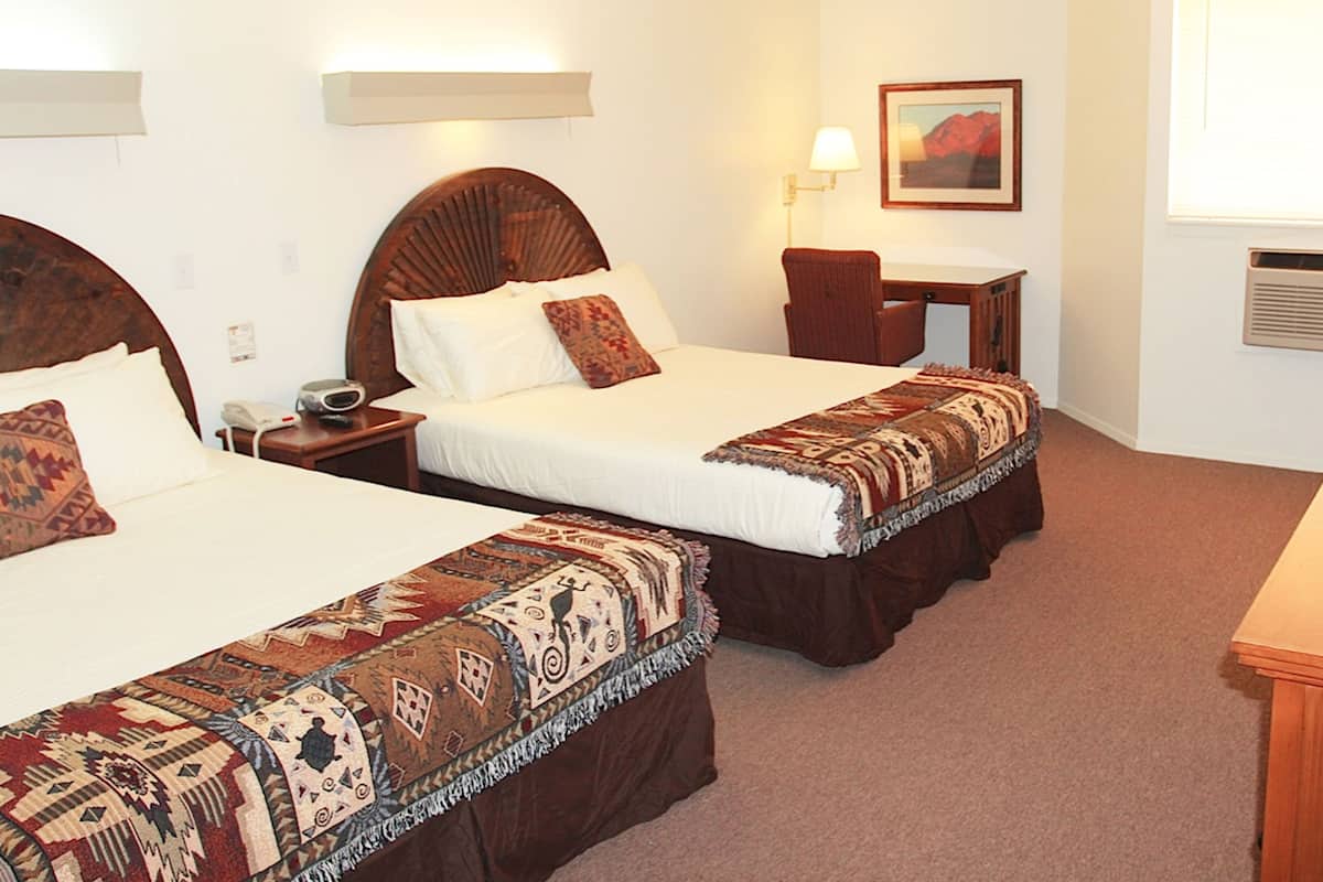 A hotel room with two beds, a desk, a chair, a lamp, and a painting on the wall.