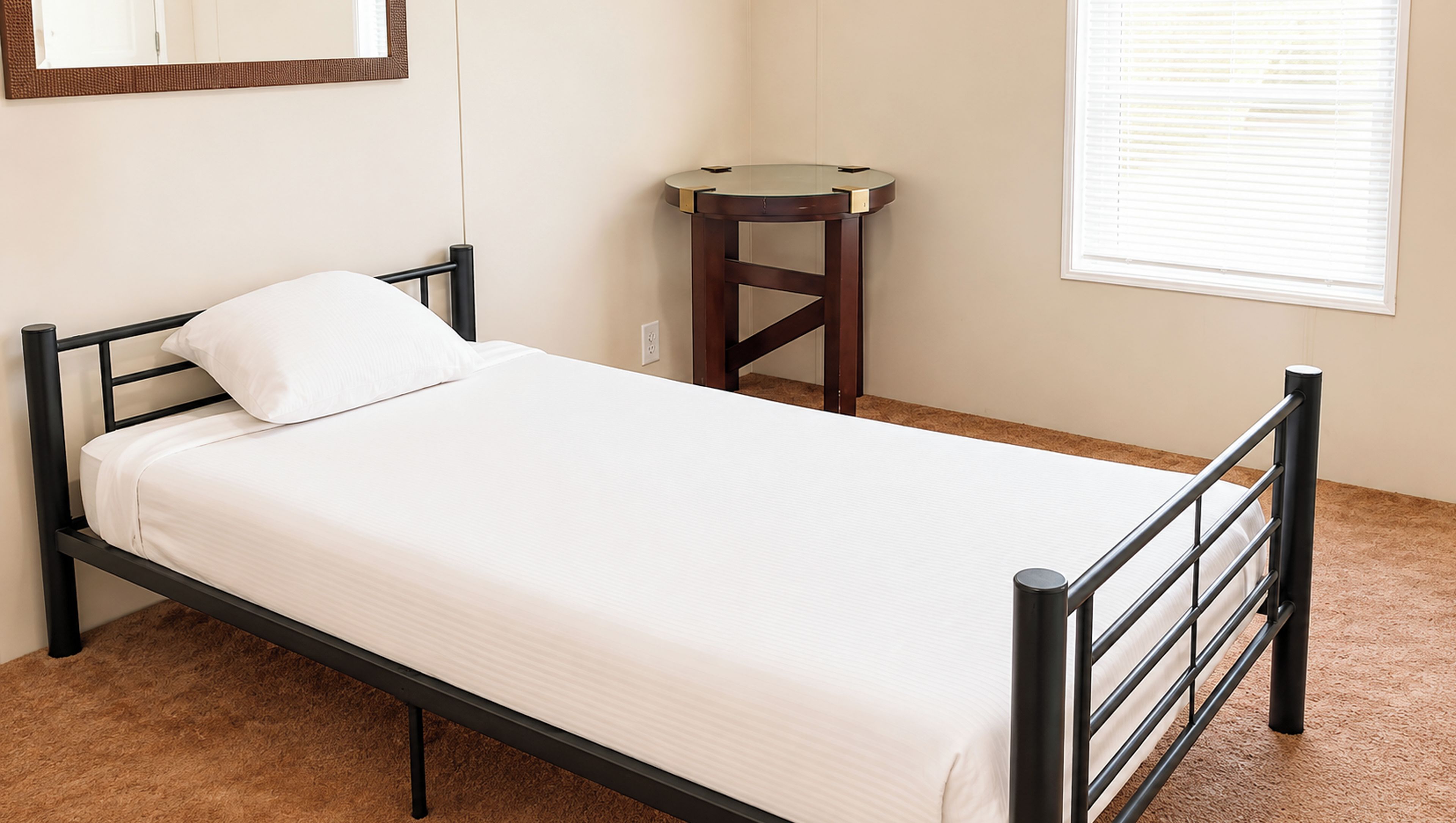 A single bed with a wooden frame and white bedding in a room with a small round table and a window with blinds.
