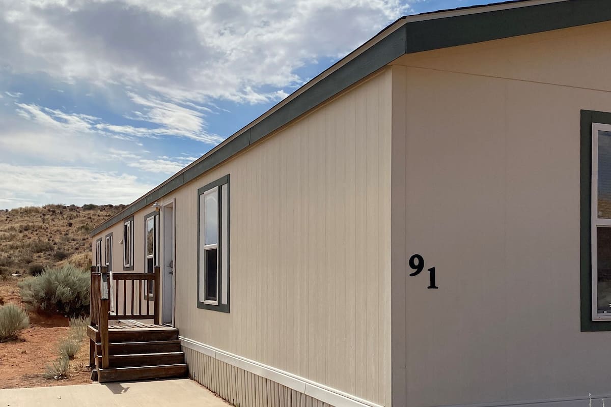 A single-story mobile home with a number '91' on the side, a front porch with steps, and a window with a view of the surrounding landscape.