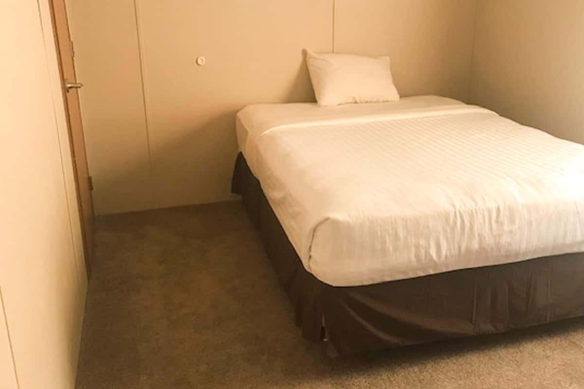 A small, single-bed room with a plain white bedspread, a dark-colored mattress, and a beige carpet.