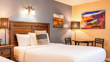 A hotel room with a wooden headboard, a bed with white bedding, two wall-mounted lamps, a small table with chairs, and a large landscape painting on the wall.