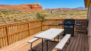 Una terraza de madera con mesa de picnic y parrilla, con vista a un paisaje desértico y formaciones rocosas rojas.
