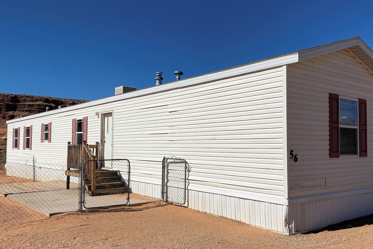 A single-story, white mobile home with a small porch, a door, and a window with shutters.