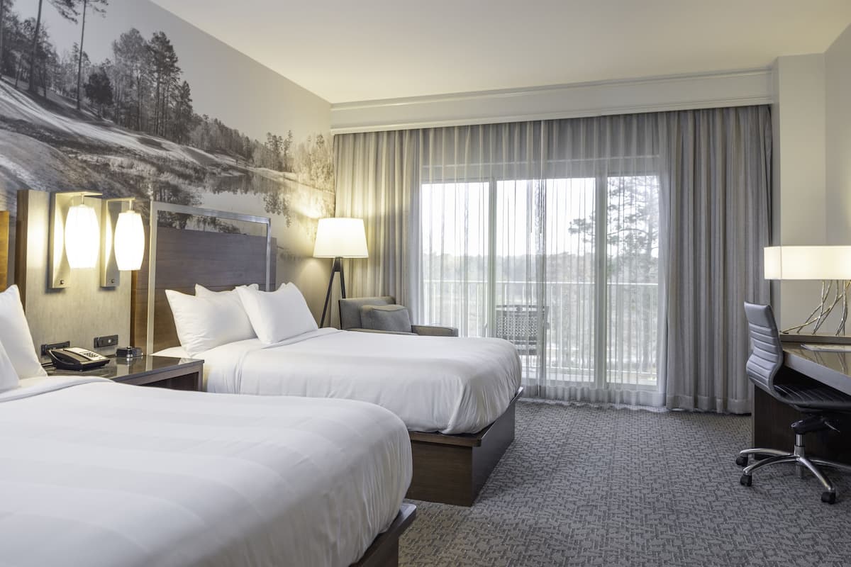 Ein Hotelzimmer mit zwei Betten, einem Schreibtisch, einem Sessel, einem Fernseher und einem großen Fenster mit Vorhängen.