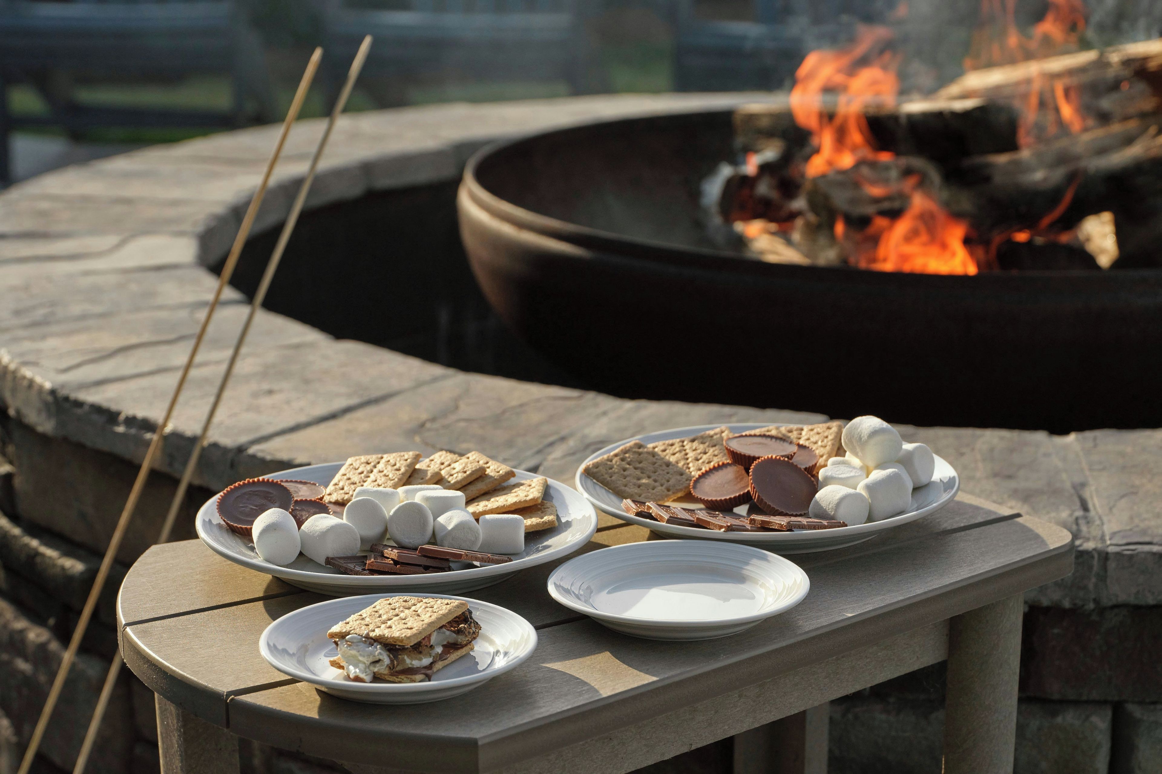 A stone fire pit with a fire burning, surrounded by a stone bench with plates of s'mores and marshmallows.