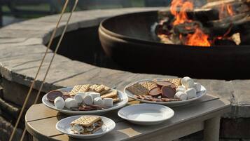 A stone fire pit with a fire burning, surrounded by a stone bench with plates of s'mores and marshmallows.