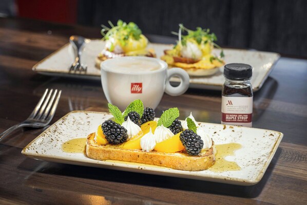 A breakfast setting with toast topped with fruit and whipped cream, a coffee cup, and a bottle of maple syrup.