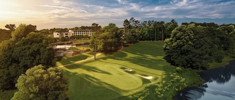 A golf course with a water body on one side, surrounded by dense trees and a building in the background.