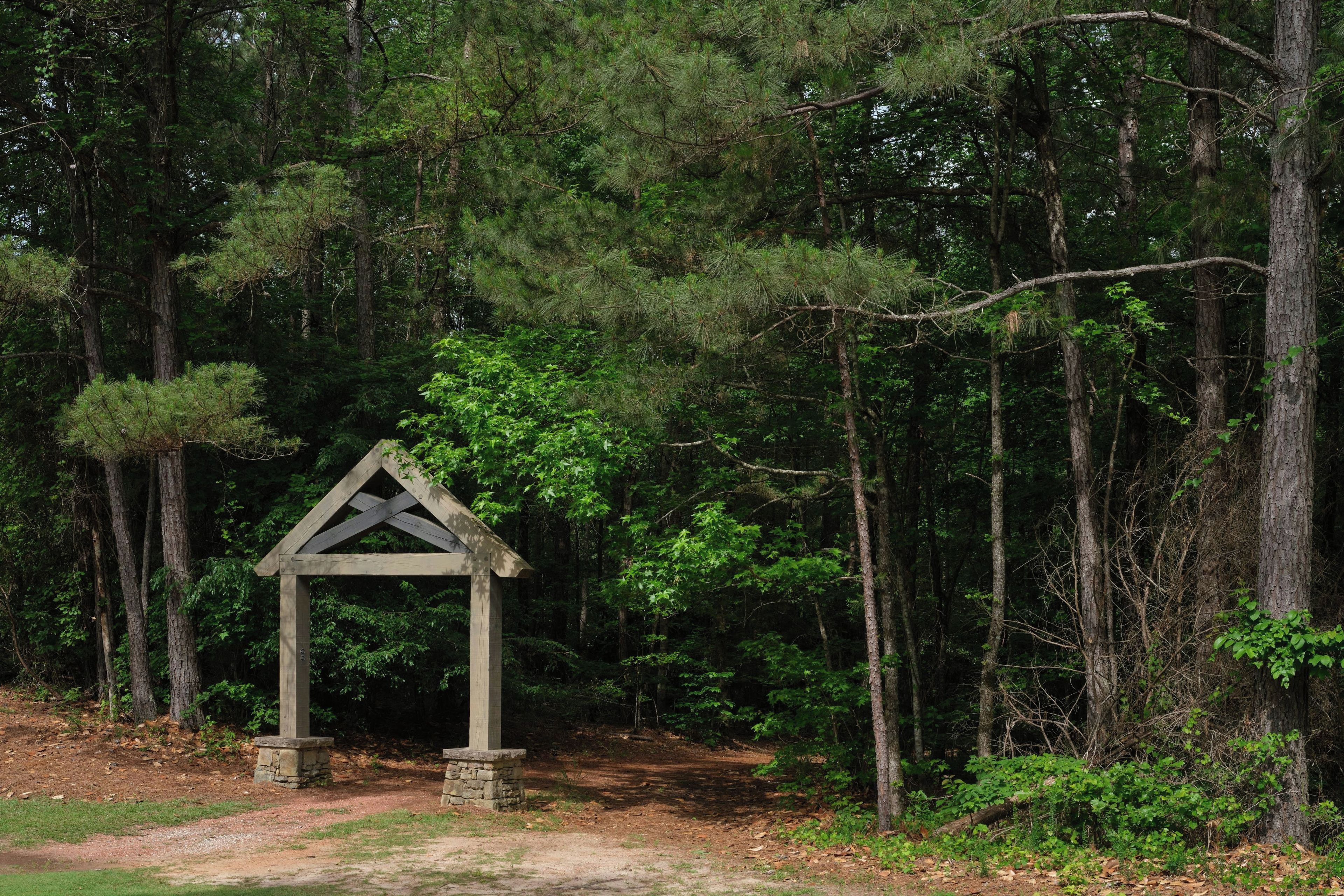 A wooden gazebo in a forested area with a dirt path leading to it.