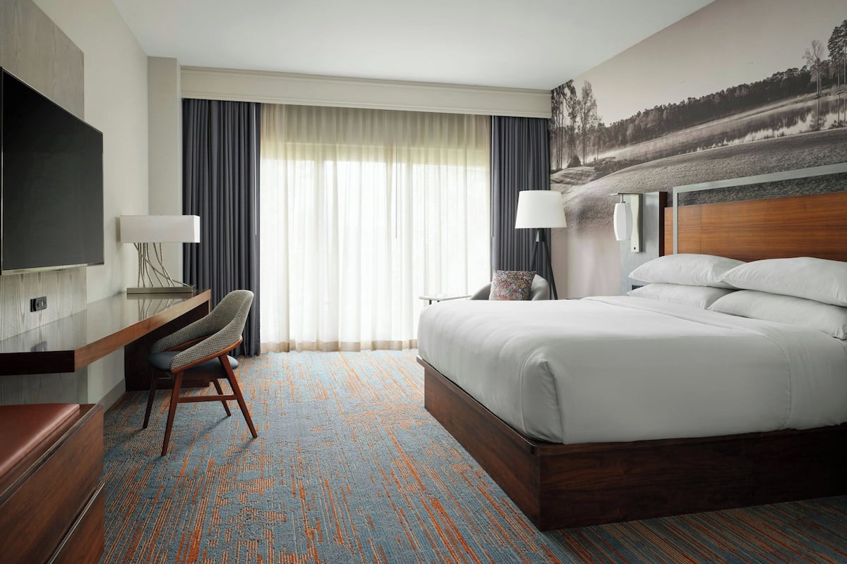Ein Hotelzimmer mit einem Bett, einem Schreibtisch, einem Sessel, einem Fernseher und einem großen Fenster mit Vorhängen.