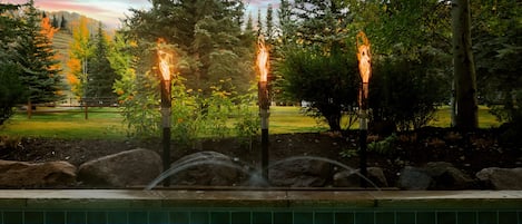 A pool area with a fountain and three torches in a landscaped garden.