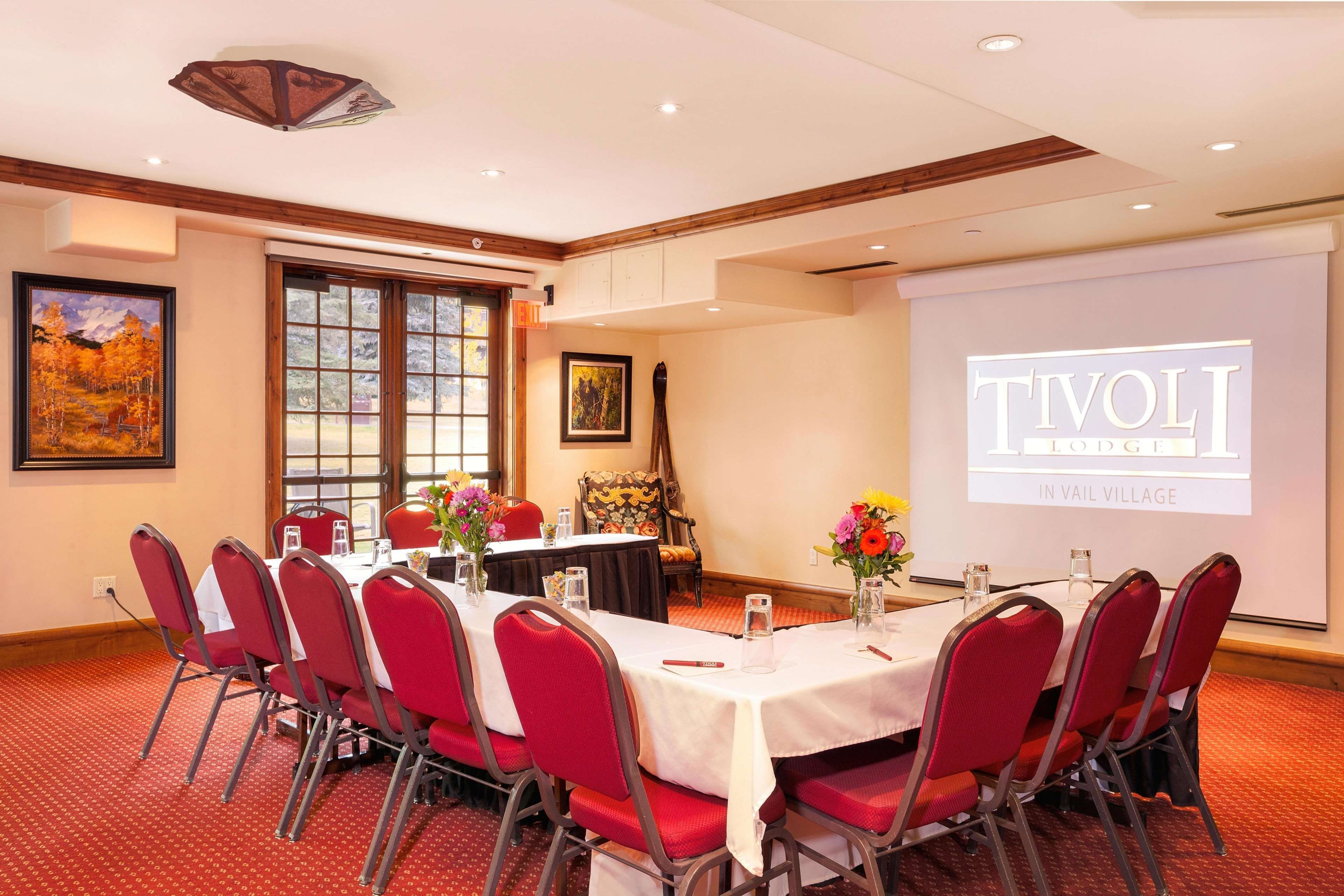 A conference room with a projection screen displaying 'TIVOLI IN VAIL VILLAGE', red chairs, a table with a white tablecloth, and framed artwork on the walls.
