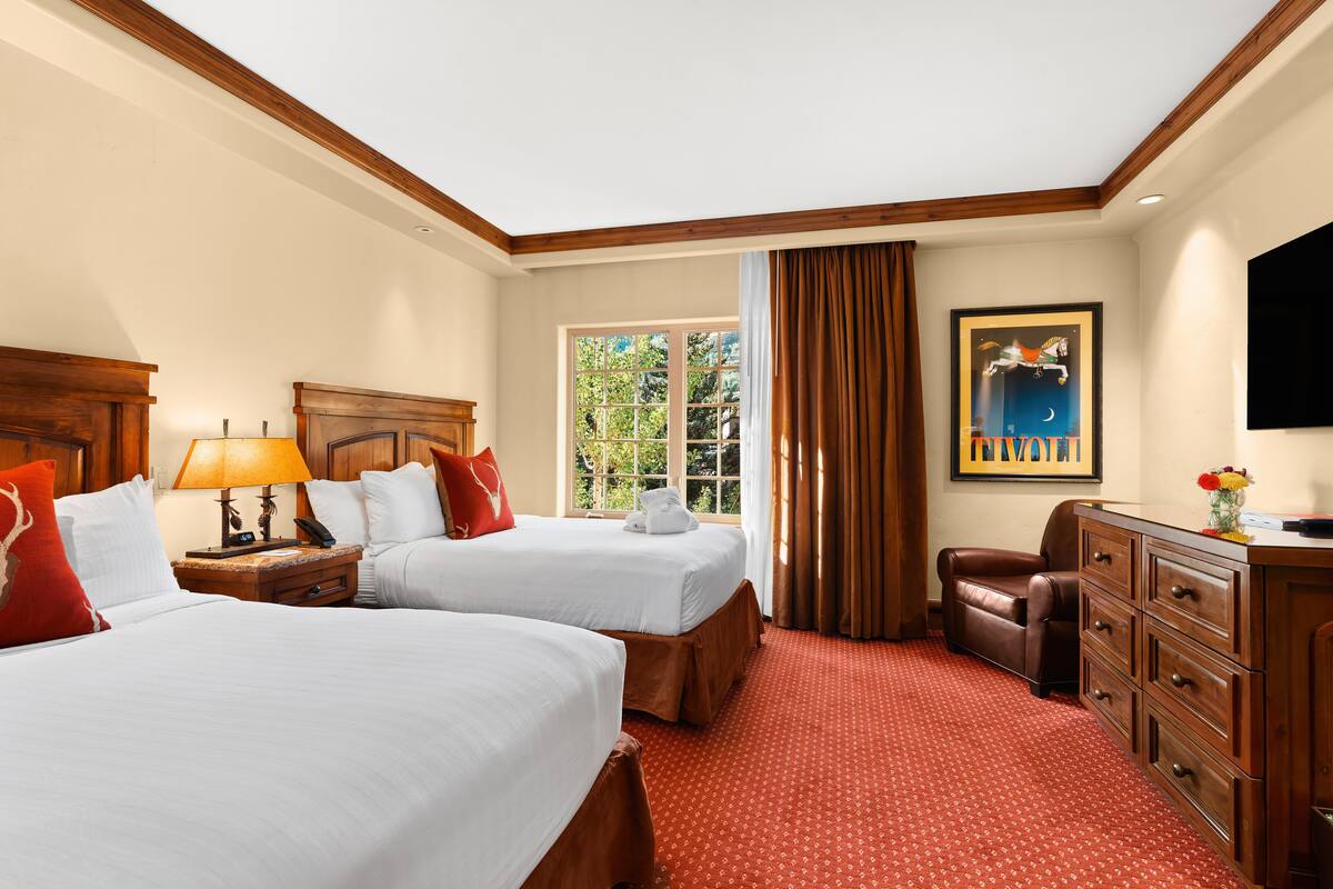 A hotel room with two beds, a wooden headboard, a desk with drawers, a television, and a window with curtains.