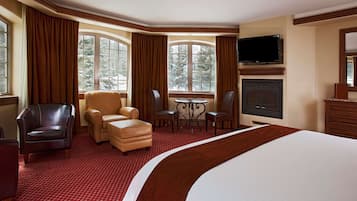 Una habitación de hotel con una cama grande, zona de estar con una mesa, chimenea y ventanas amplias con cortinas.