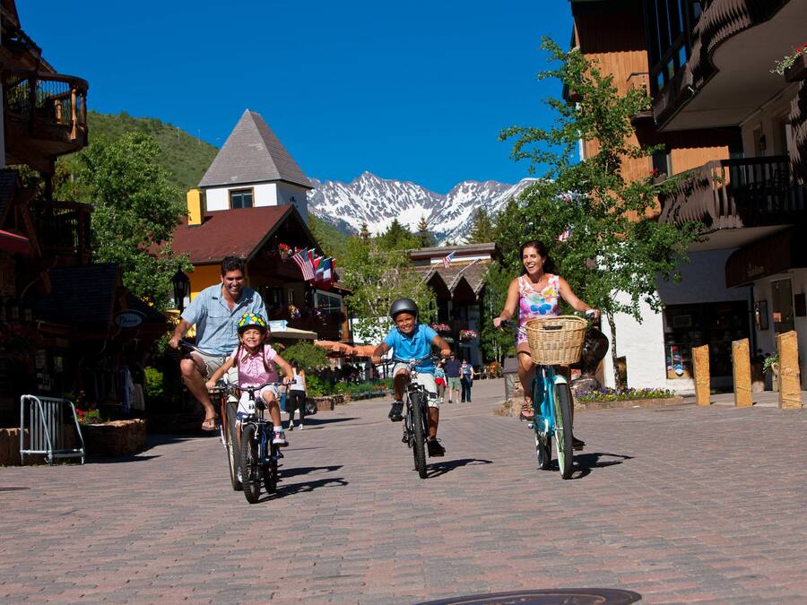 A group of people riding bicycles on a paved street with buildings and mountains in the background.