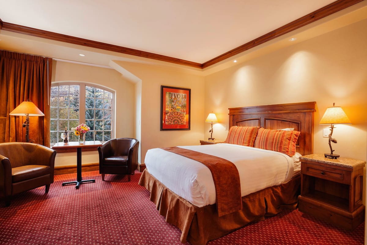 A hotel room with a large bed, two bedside lamps, a wooden headboard, a window with curtains, a desk with a chair, and a framed picture on the wall.