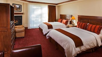 A hotel room with two beds, a fireplace, a desk with a lamp, and a leather armchair.