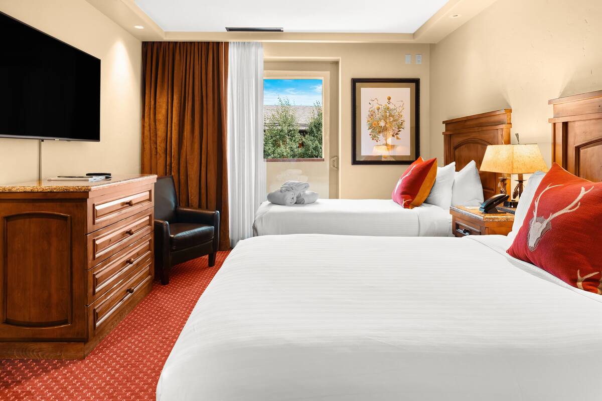 A hotel room with two beds, a TV, a desk, and a window with a view of greenery.