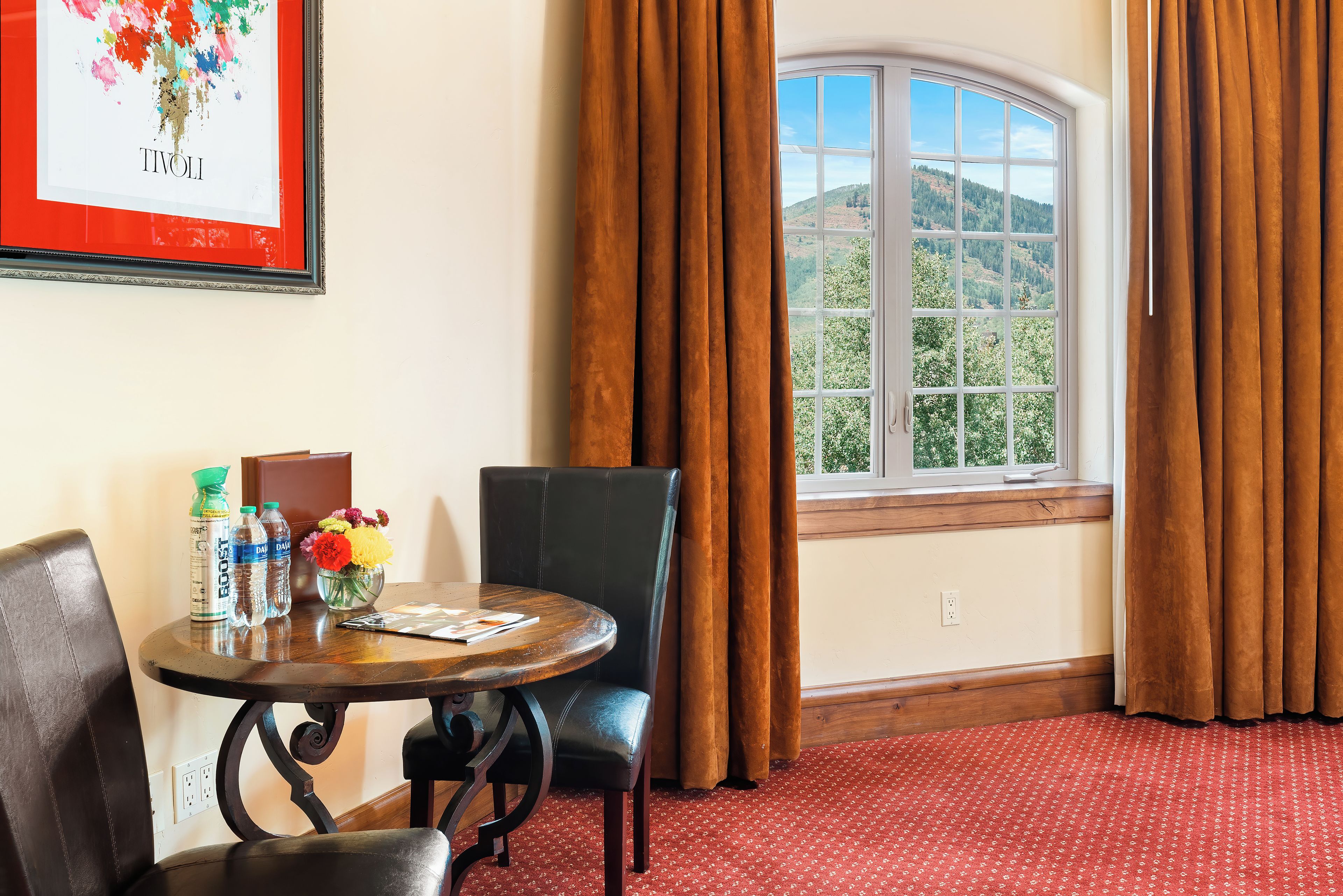 Une chambre d’hôtel avec une table à manger, des chaises, un vase de fleurs, un tableau encadré au mur et une fenêtre avec des rideaux.