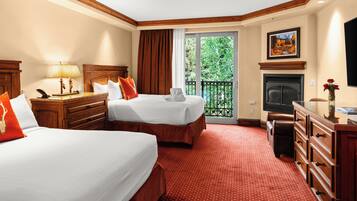 A hotel room with two beds, a desk, a dresser, a fireplace, and a balcony with a view of trees.