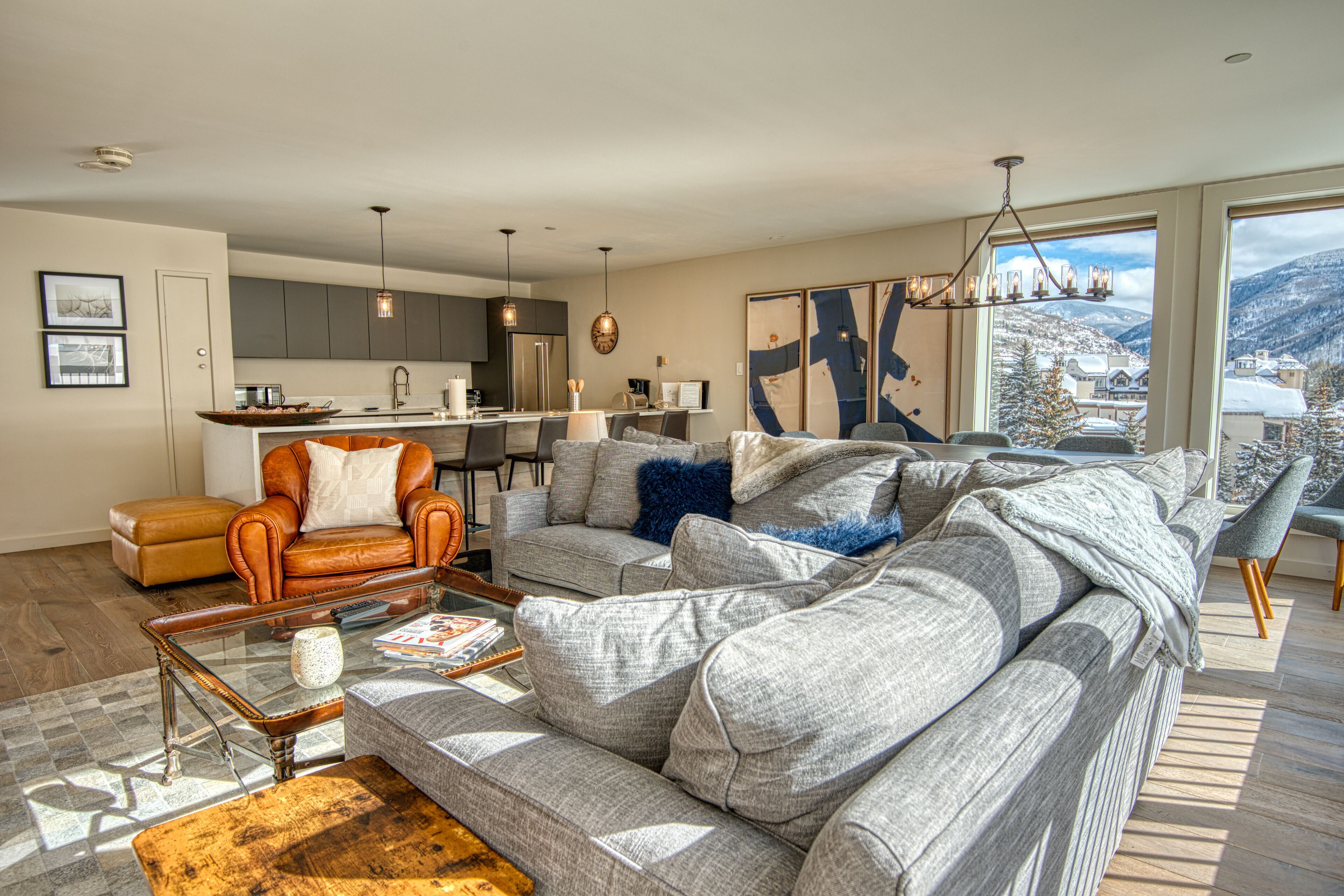 A modern living room with a grey sofa, a leather armchair, and a coffee table. There is a kitchen area in the background with a refrigerator and a dining table.