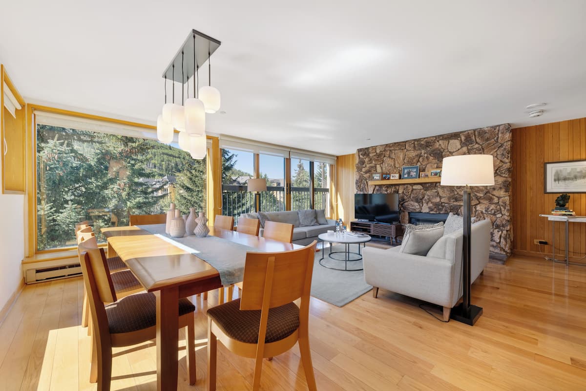 A modern living room with a dining area, large windows, a stone wall, and a fireplace.