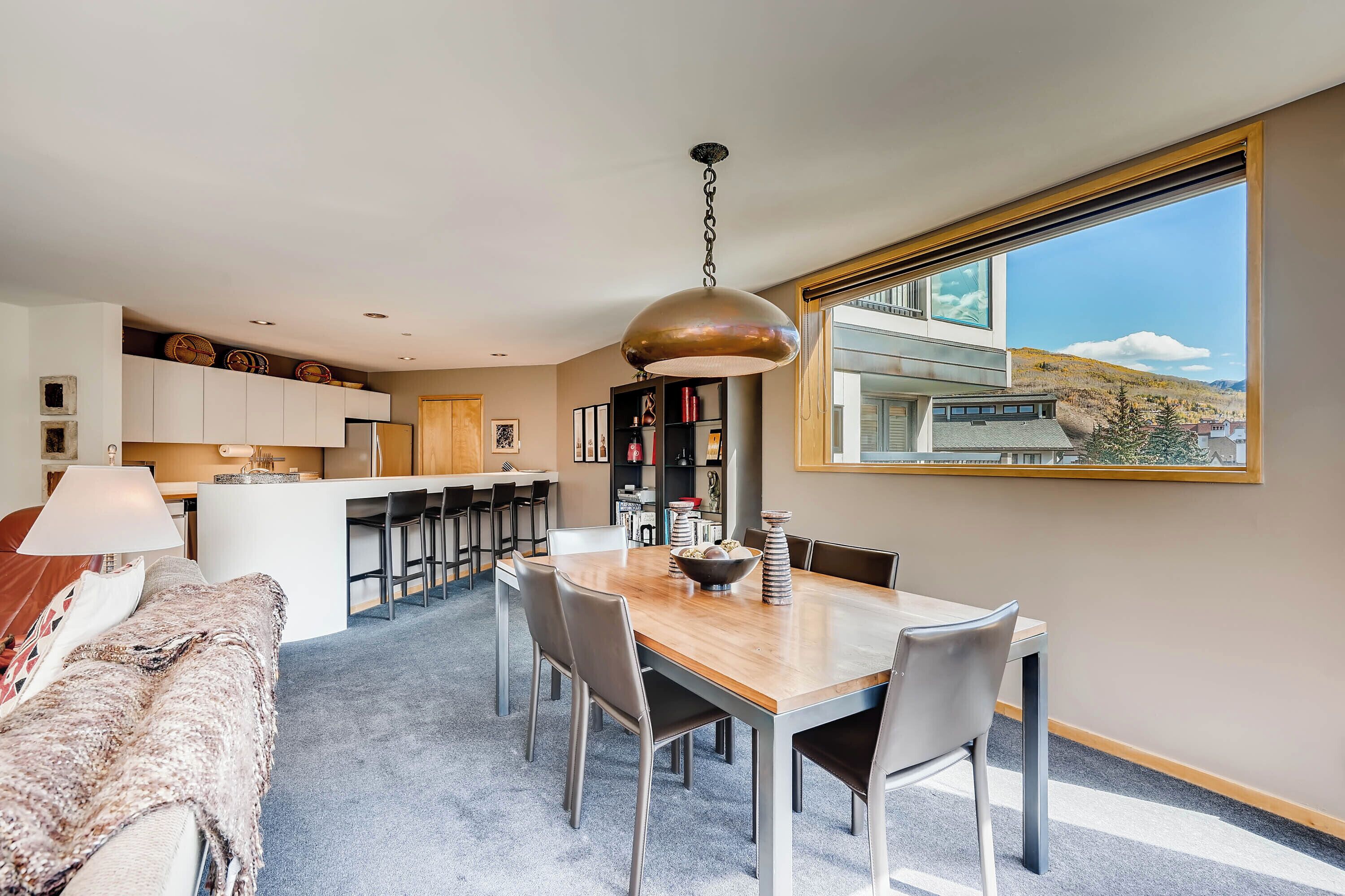 Un comedor moderno con una mesa y sillas de madera, una lámpara colgante y vistas a un paisaje montañoso a través de un ventanal grande.