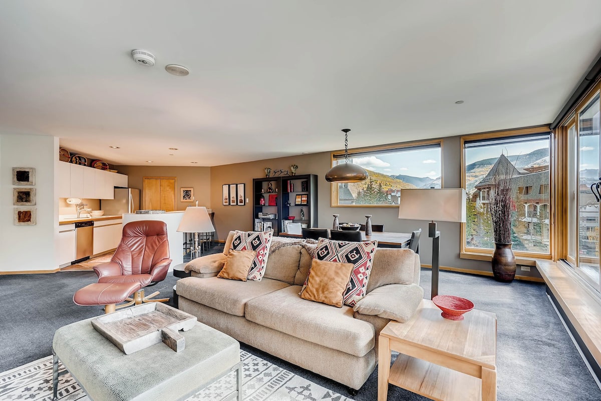 A modern living room with a sofa, coffee table, and a view of the mountains through large windows.