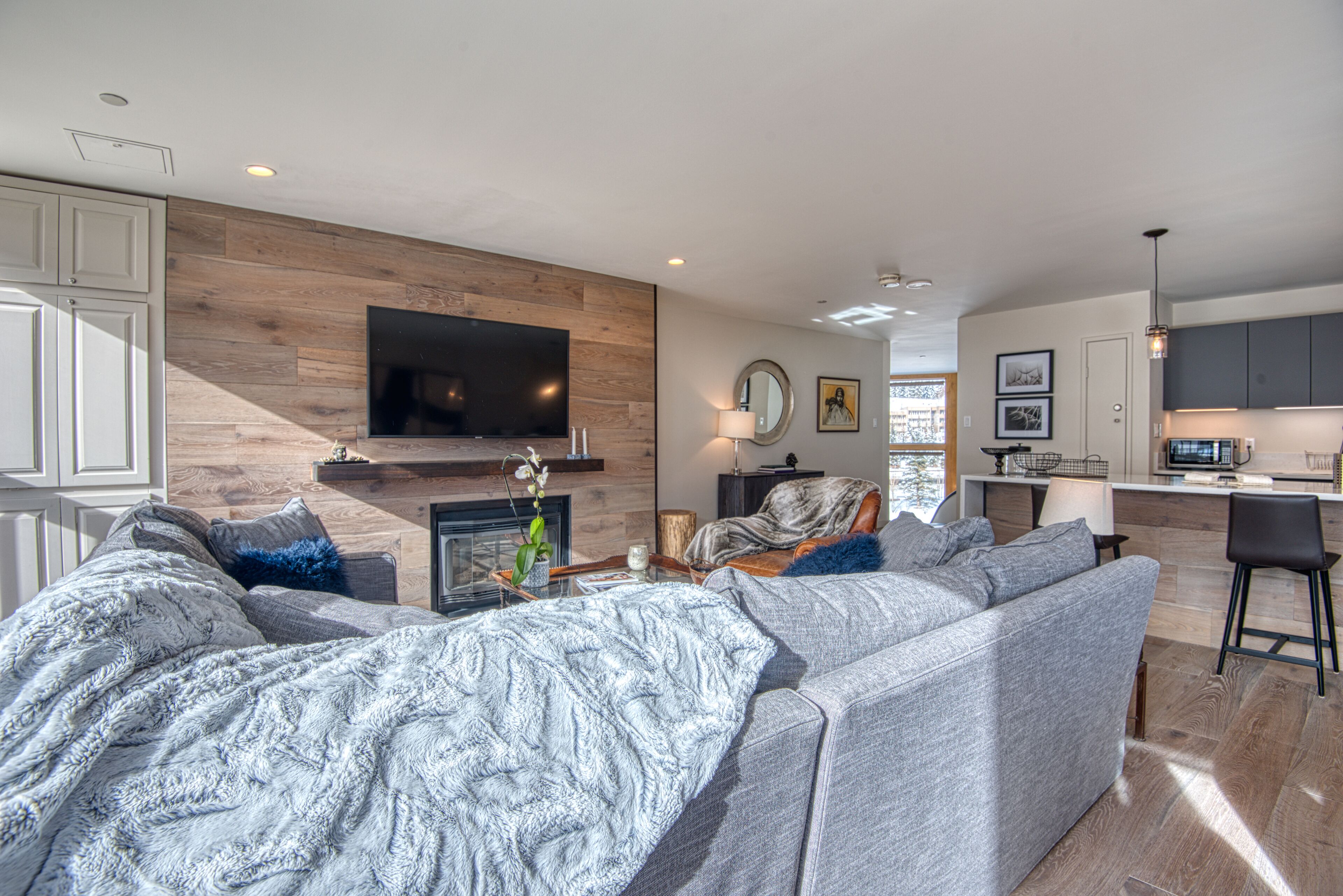 A modern living room with a grey sectional sofa, a flat-screen TV mounted on a wooden wall, and a kitchen area in the background.