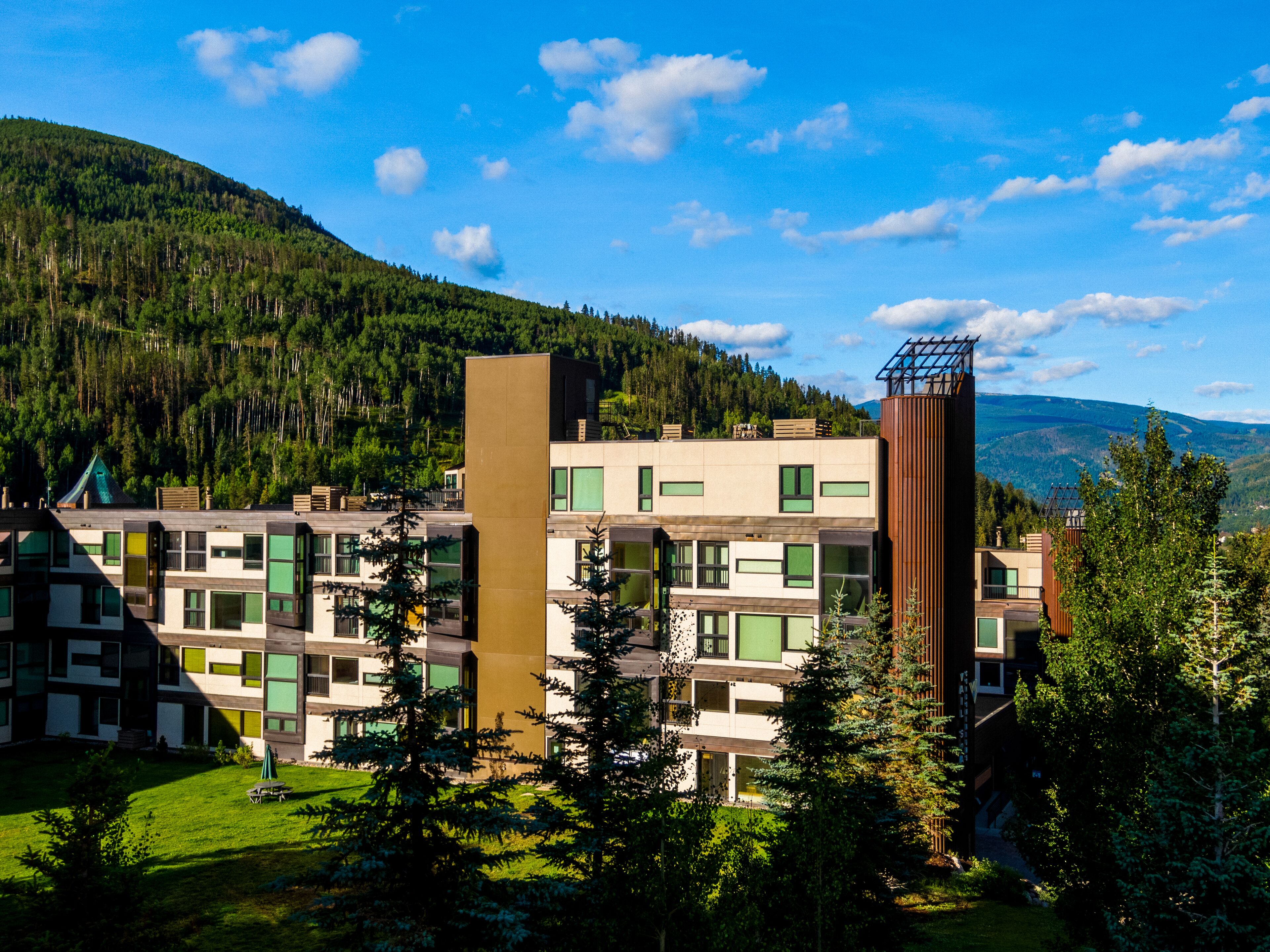 A modern multi-story building with a flat roof and large windows, surrounded by lush greenery and mountains in the background.