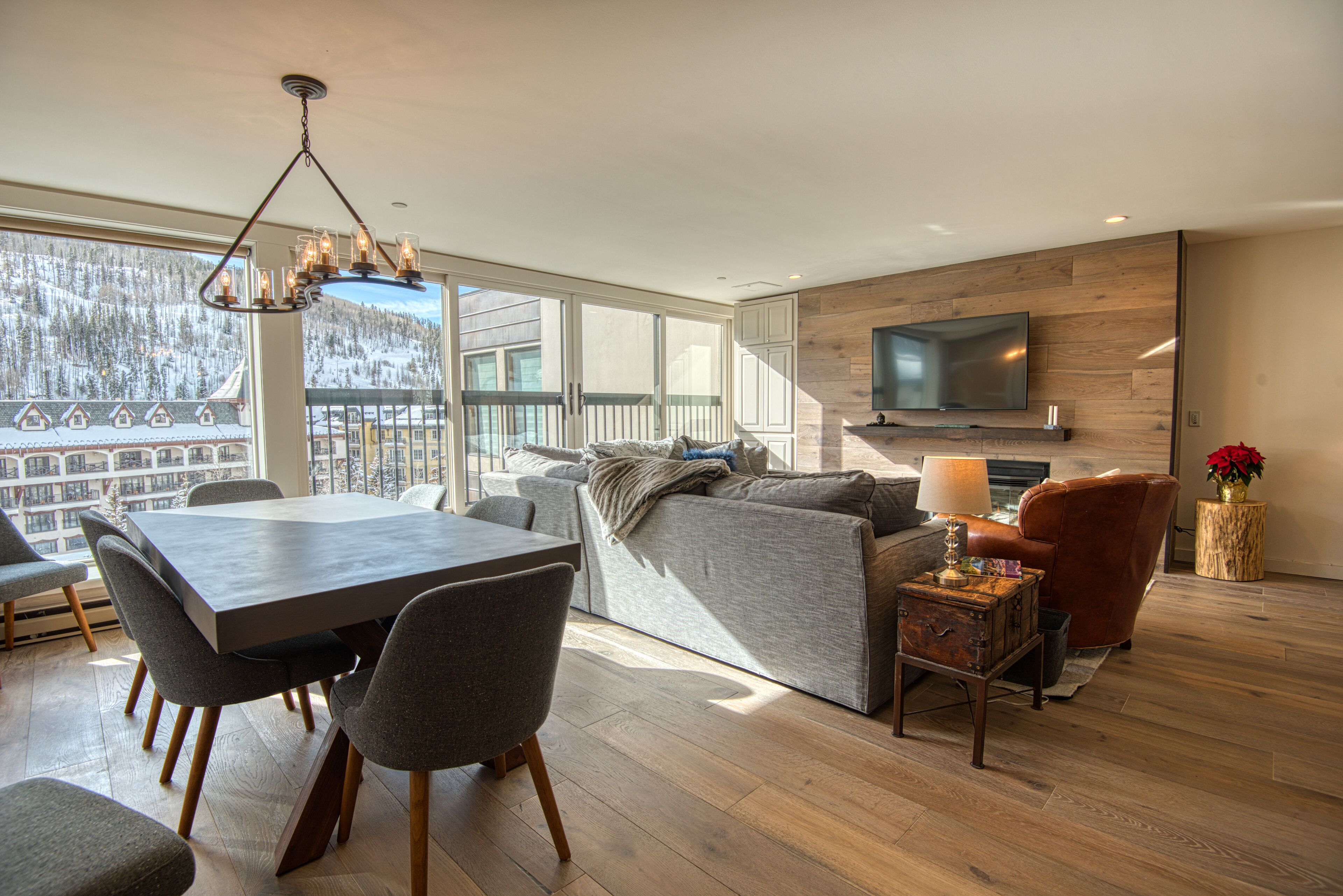 A modern living room with a dining area, large windows, a fireplace, and a TV mounted on the wall.