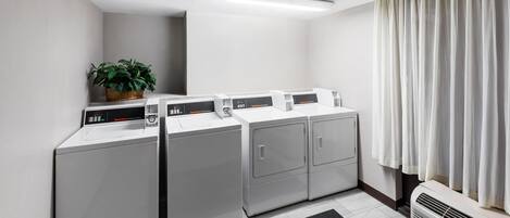 A laundry room with four front-loading washers and dryers, a potted plant, and a large window with curtains.