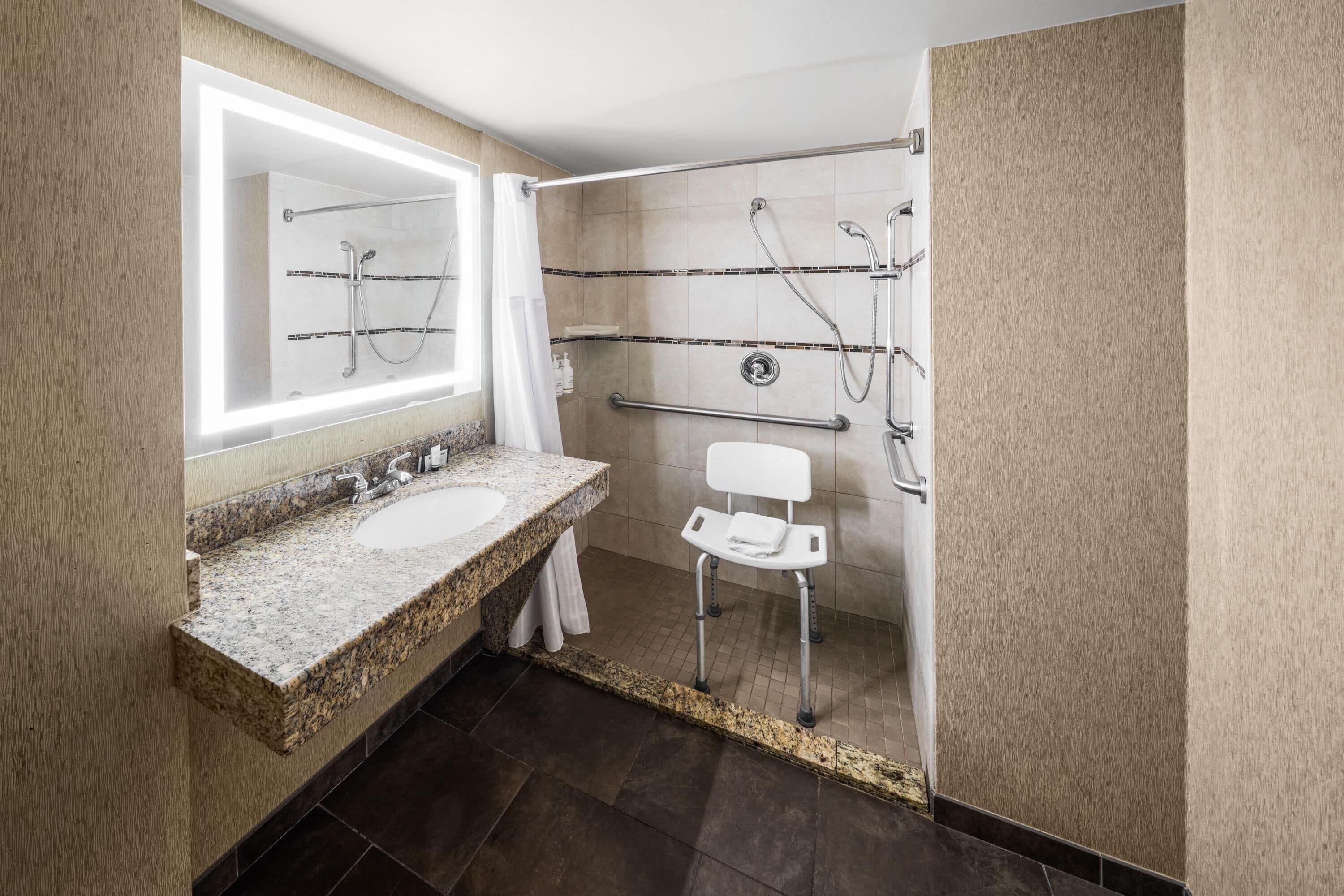 A modern bathroom with a granite countertop sink, a mirror, a shower with a handheld showerhead, and a grab bar.