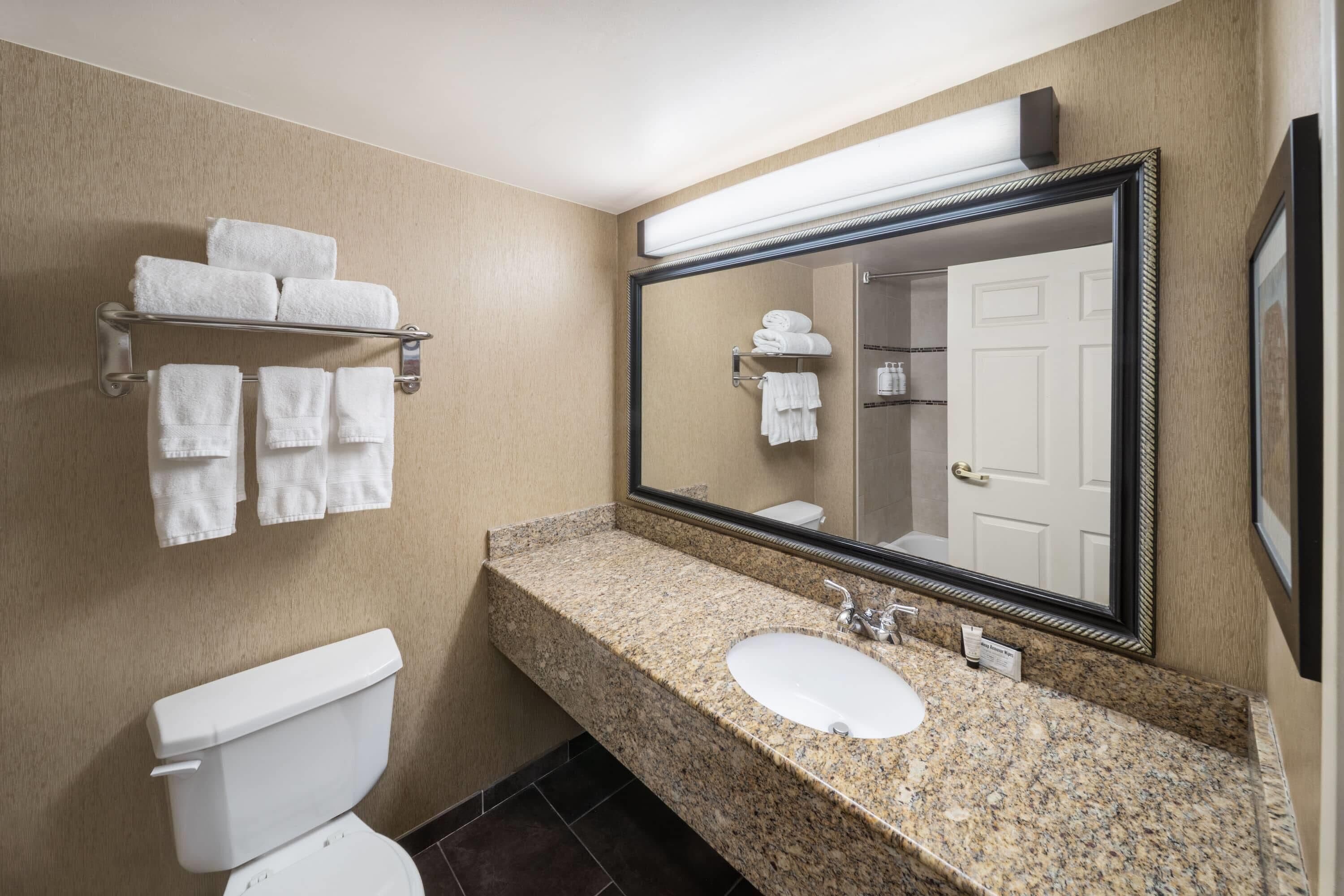 A bathroom with a granite countertop, a white sink, a toilet, and a mirror with a towel rack above it.