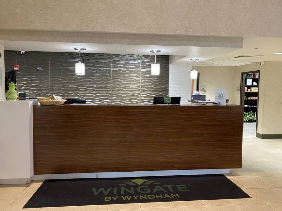 A reception desk with a wooden finish, a black mat with "Wingate by Wyndham" logo, and a textured wall behind.