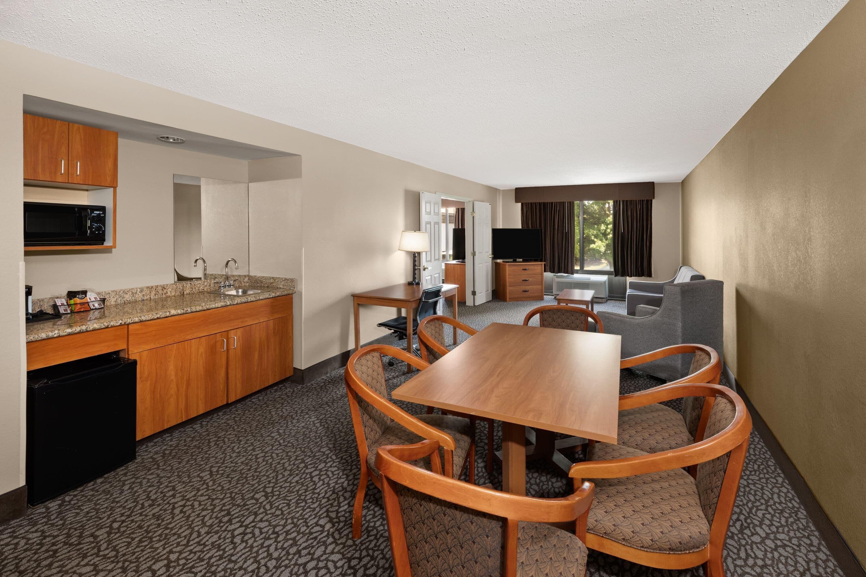 A hotel room with a kitchenette, a dining area with a table and chairs, a sofa, a TV, and a window with curtains.