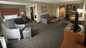 A hotel room with a bed, grey sofa, armchair, small table, TV, and kitchenette. - Wingate by Wyndham Little Rock (Little Rock)