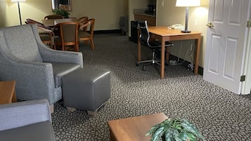 A hotel room with a grey sofa, a wooden coffee table with a plant, a dining table with chairs, a small kitchen area, and a door.