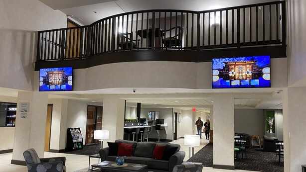 A spacious lobby with a curved balcony, a sofa set, and two televisions displaying different scenes. - Wingate by Wyndham Little Rock (Little Rock)
