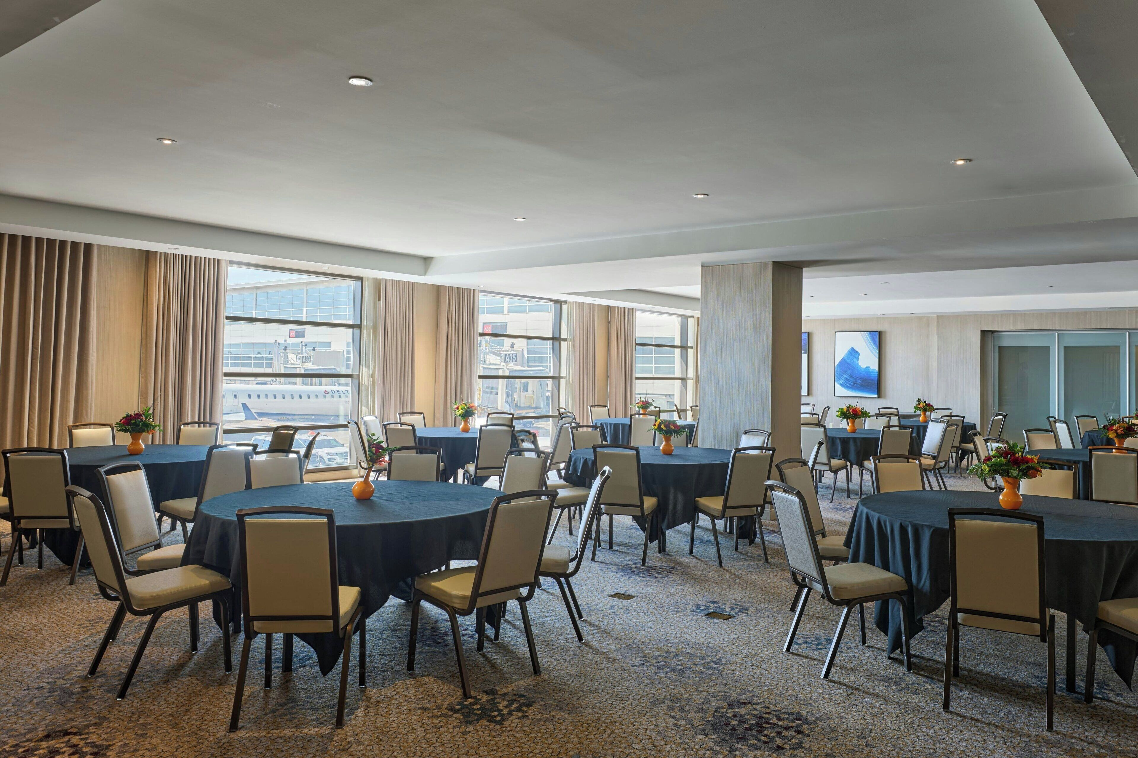 A large conference room with multiple round tables and chairs, large windows with curtains, and a city view.