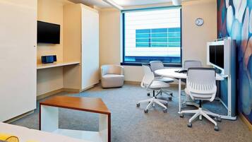 A modern office space with a flat-screen TV, a round table with chairs, a sofa, and a large window with blinds.