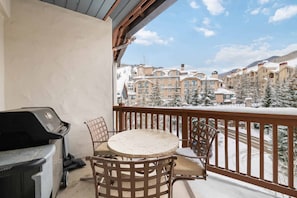 Un balcón con una parrilla y sillas, con vista a un pueblo cubierto de nieve, con edificios y árboles.