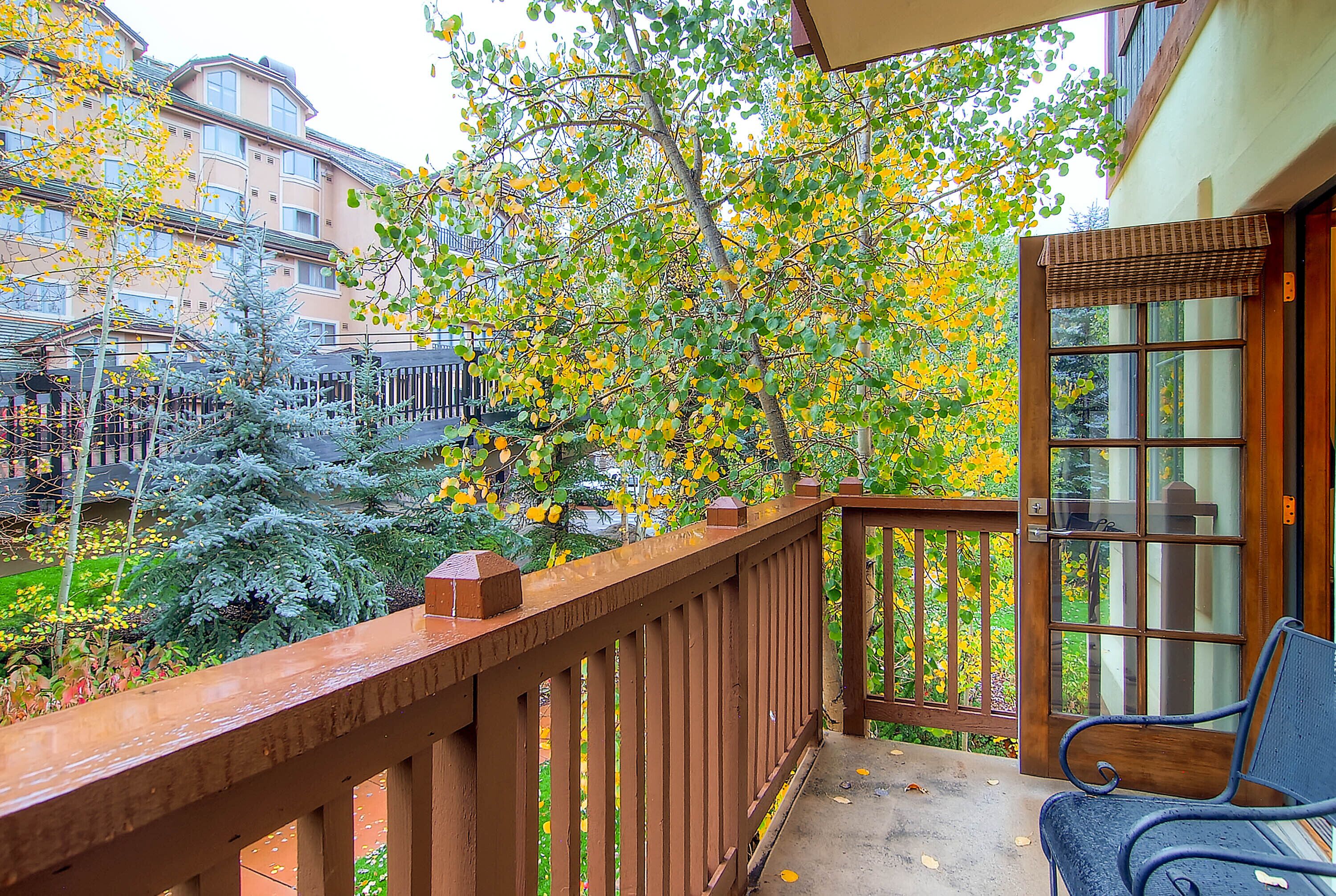 A balcony with a wooden railing, a bench, and a view of a building with balconies and trees with yellow leaves.