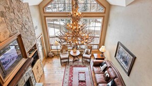 A cozy living room with a stone fireplace, leather sofa, and a large window with a view of snow-covered trees. - Oxford Court by East West Hospitality (Avon)