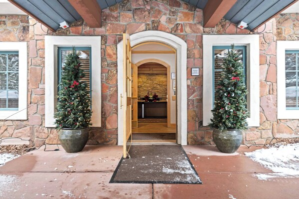 Entrada de un edificio de piedra con una puerta de madera, dos árboles de Navidad en macetas y un felpudo.