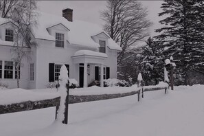 A snow-covered house with a chimney, windows, and a porch. - 1824 House (Waitsfield)
