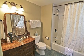 A bathroom with a wooden vanity, a large mirror, a glass bowl sink, a toilet, and a walk-in shower with a patterned curtain. - 1824 House (Waitsfield)