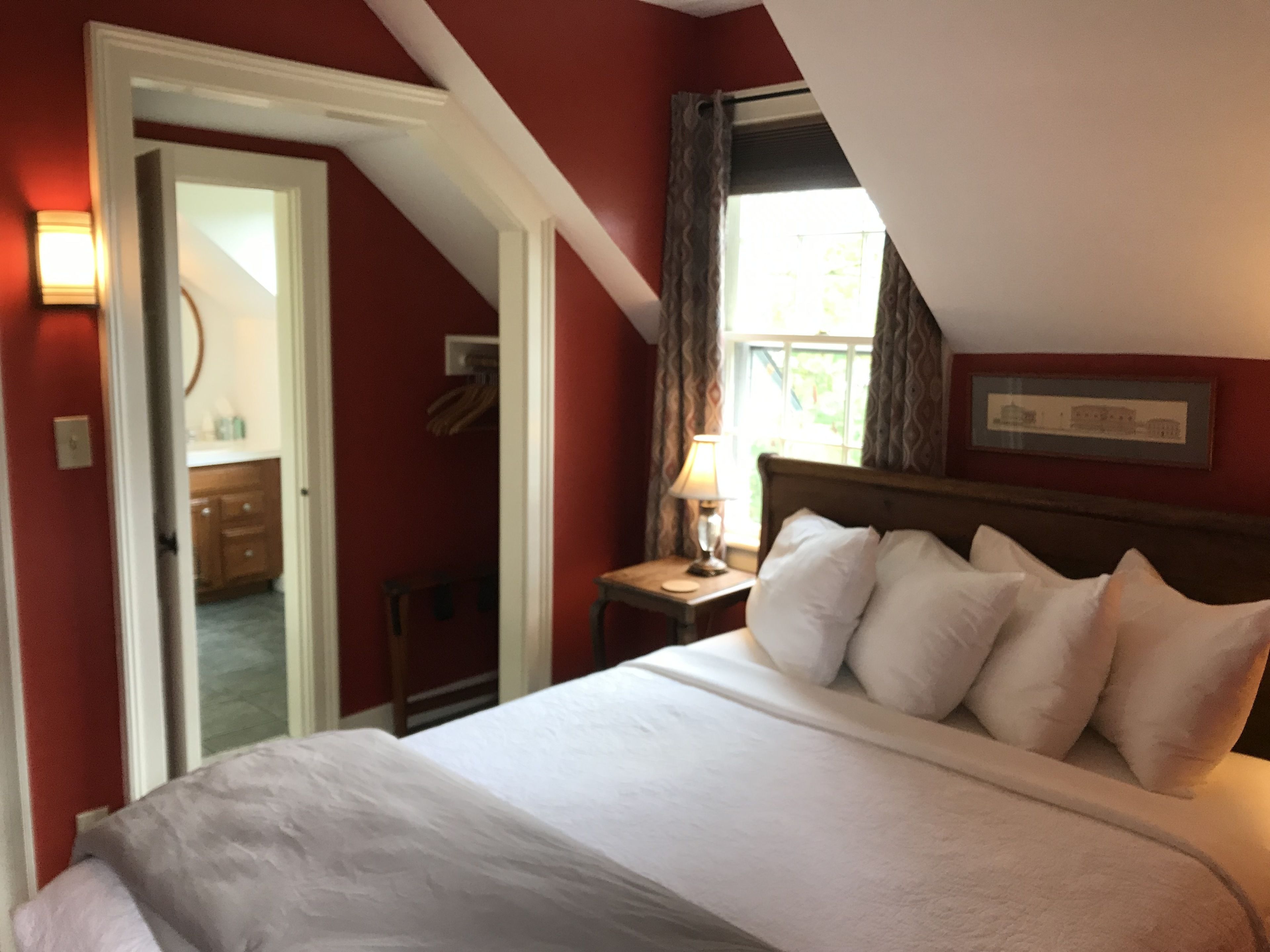 Un dormitorio con una cama grande, una mesita de noche con lámpara, una ventana con cortinas y una puerta que da a otra habitación.