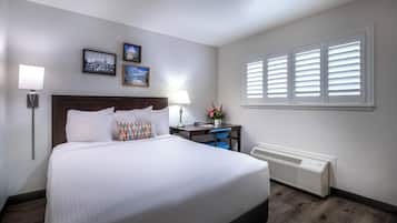 A hotel room with a large bed, a desk, a chair, a desk lamp, a wall-mounted heater, and a window with blinds.