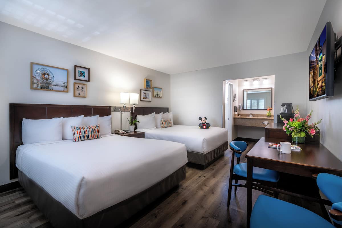 Ein Hotelzimmer mit zwei Betten, einem Schreibtisch, einem Stuhl und einem durchgehenden Türbogen einsehbarer Badezimmer.